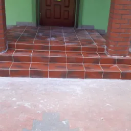 Wykończone schody wejściowe z czerwonobrązowych płytek ceramicznych z białymi fugami, prowadzące do drzwi wejściowych, z kolumnami obłożonymi cegłą po bokach, ślady białego pyłu na płytkach i podłożu.