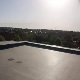 Płaski dach pokryty papą termozgrzewalną na tle zabudowy miejskiej w słoneczny dzień, widoczne ślady po naprawach.