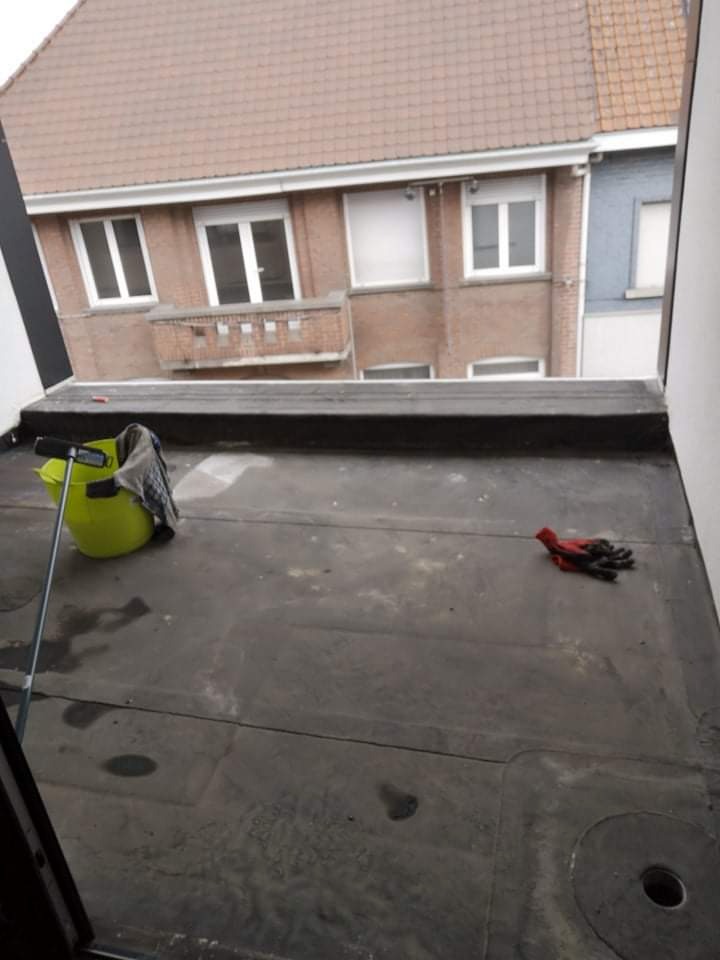 Płaski dach pokryty papą, widoczny z góry z fragmentem budynku w tle, z wiaderkiem i rękawicami roboczymi na powierzchni.