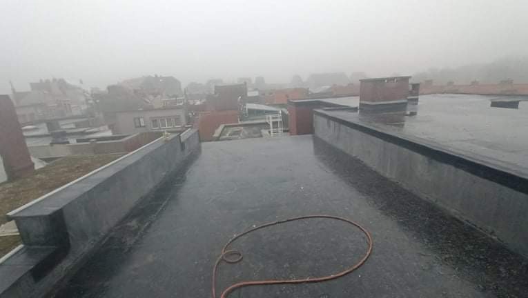 Płaski dach pokryty czarną papą w deszczowy dzień, widoczne kominy i budynki w tle, zwinięty wąż leżący na powierzchni.