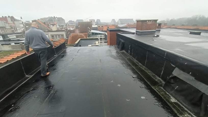 Czarny, płaski dach pokryty papą bitumiczną, z widocznymi obróbkami blacharskimi i stojącym dekarzem w szarym swetrze, na tle zabudowy miejskiej w pochmurny dzień.