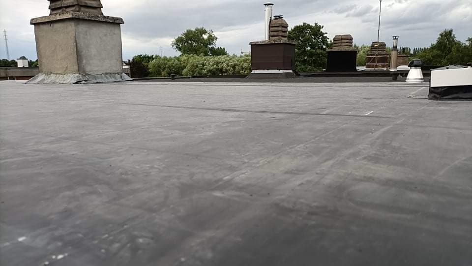Płaski dach pokryty czarną membraną bitumiczną, widoczne kominy i elementy wentylacyjne na tle zieleni i pochmurnego nieba.