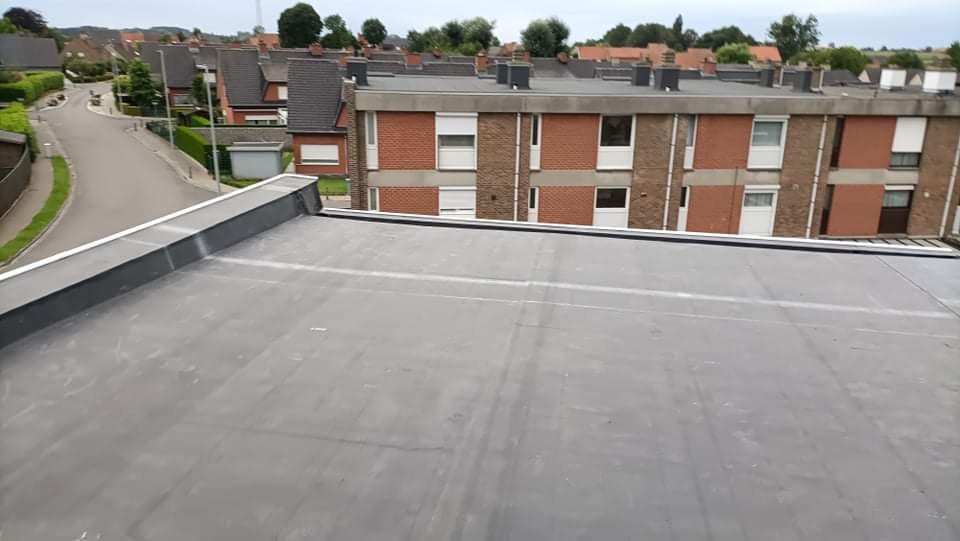Widok z góry na płaski dach pokryty papą, z widocznymi budynkami mieszkalnymi w tle i elementami obróbki blacharskiej na krawędzi dachu.