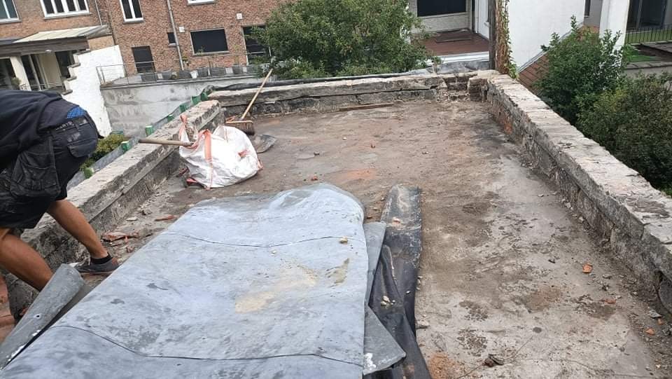 Płaski dach w trakcie renowacji, widoczne stare pokrycie, gruz, torba z odpadami i fragment sylwetki pracownika.