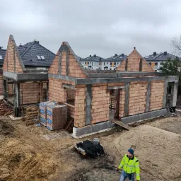 Wznoszenie ścian parterowego domu z czerwonej cegły, widok z góry, z pracownikiem w odblaskowej kurtce na tle placu budowy i sterty piasku.