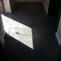 Ułożona, ciemnoszara podłoga z płytek w korytarzu, widziana z perspektywy wejścia, z wyraźnym cieniem rzucanym przez nieznany obiekt.