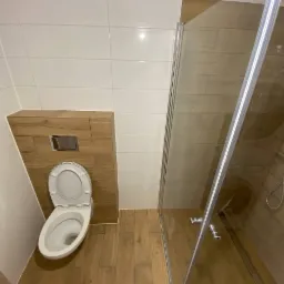 Nowoczesna łazienka z drewnopodobnymi płytkami na podłodze i ścianie, białe płytki na górze, toaleta podwieszana z zabudową drewnopodobną, kabina prysznicowa z transparentnego szkła.