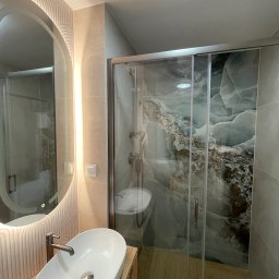 Welcome Home - Nowoczesna łazienka z owalnym lustrem z podświetleniem LED, umywalką nablatową i kabiną prysznicową z płytkami imitującymi marmur. Drewniane wykończenie szafki pod umywalką.