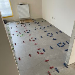 Welcome Home - Szare płytki podłogowe w trakcie układania, z użyciem kolorowych klinów poziomujących. Widoczny grzejnik, okno z folią ochronną i fragment ściany z gniazdkami.