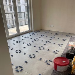 Welcome Home - Jasne płytki podłogowe w trakcie układania, z użyciem niebieskich krzyżyków dystansowych. Widoczne wiadro z klejem i narzędzia. W tle okno z widokiem na budynek.