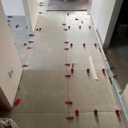 Welcome Home - Szare płytki podłogowe w trakcie układania, z czerwonymi i niebieskimi klipsami poziomującymi. Widoczne worki z zaprawą i poziomica. Prace glazurnicze w korytarzu.