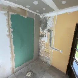 Wnętrze w trakcie prac: fragment ściany z zieloną płytą kartonowo-gipsową, surowy mur z pozostałościami tynku obok, oraz fragment pomalowanej ściany z gniazdkiem elektrycznym. Widoczne ślady po...