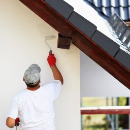 Kamil Kostka - Pracownik w czerwonej rękawicy maluje elewację budynku we Wrocławiu za pomocą wałka; widoczna drabina i fragment dachu z ciemną dachówką.