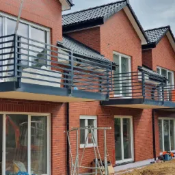 Nowoczesne, czarne balustrady balkonowe z poziomych profili zamontowane w nowym budynku z czerwonej cegły, widoczne rusztowania i sprzęt budowlany.
