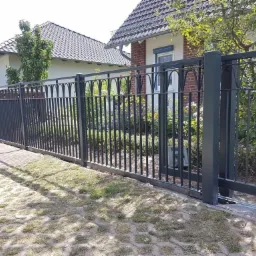 Przesuwna brama z metalowymi prętami i ozdobnymi łukami na tle domu z czerwonej cegły i zieleni ogrodu, widoczny mechanizm automatycznego otwierania.