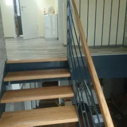 Drewniane schody z metalową balustradą o minimalistycznym designie, widok z dołu na górę, jasne wnętrze z otwartymi drzwiami w tle.