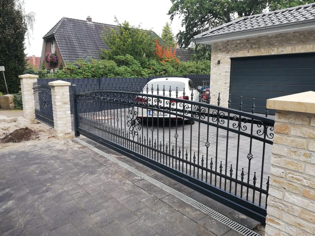 Czarna, metalowa brama wjazdowa z ozdobnymi detalami, osadzona pomiędzy murowanymi słupkami z jasnej cegły, na tle domu z garażem i samochodu osobowego.
