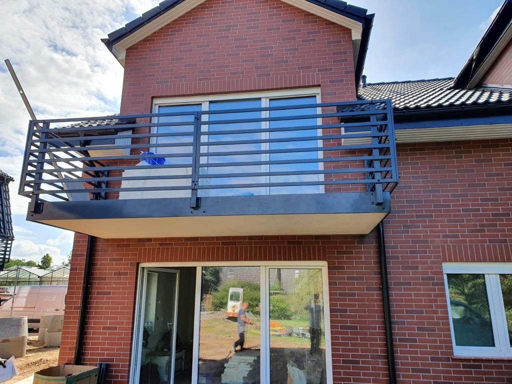 Nowoczesna, czarna balustrada balkonowa z poziomych prętów zamontowana na balkonie domu z czerwonej cegły; widoczne okna i otoczenie budynku w trakcie budowy.