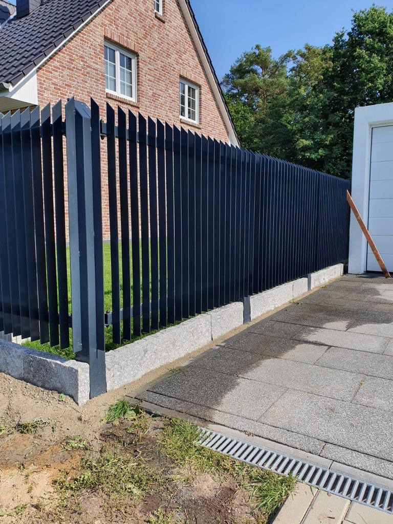 Nowoczesne, pionowe, czarne ogrodzenie z metalowych profili, osadzone na betonowej podmurówce, przed domem z czerwonej cegły.