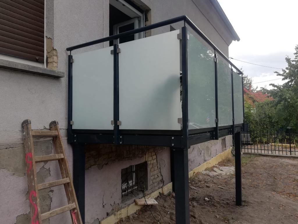 Nowoczesna balustrada balkonowa z czarną metalową ramą i matowymi szklanymi panelami, zamontowana na podwyższonym balkonie przy szarym budynku z widoczną drewnianą drabiną opartą o ścianę.