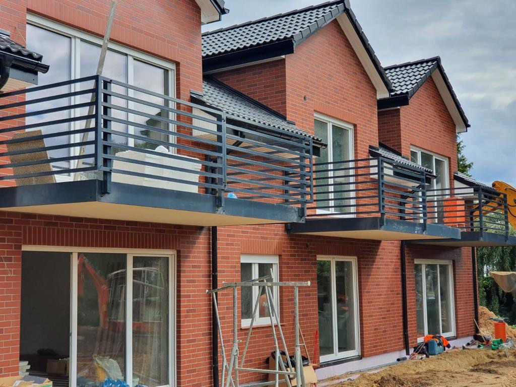 Nowoczesne, czarne balustrady balkonowe z poziomych profili zamontowane w nowym budynku z czerwonej cegły, widoczne rusztowania i sprzęt budowlany.