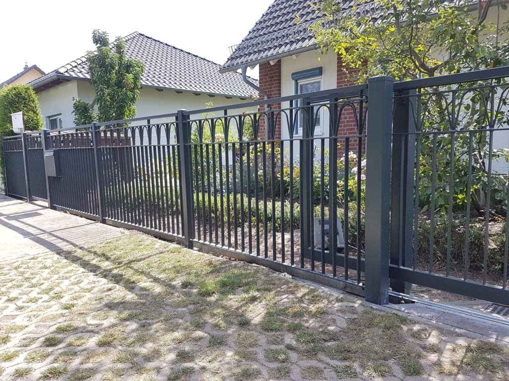 Przesuwna brama z metalowymi prętami i ozdobnymi łukami na tle domu z czerwonej cegły i zieleni ogrodu, widoczny mechanizm automatycznego otwierania.