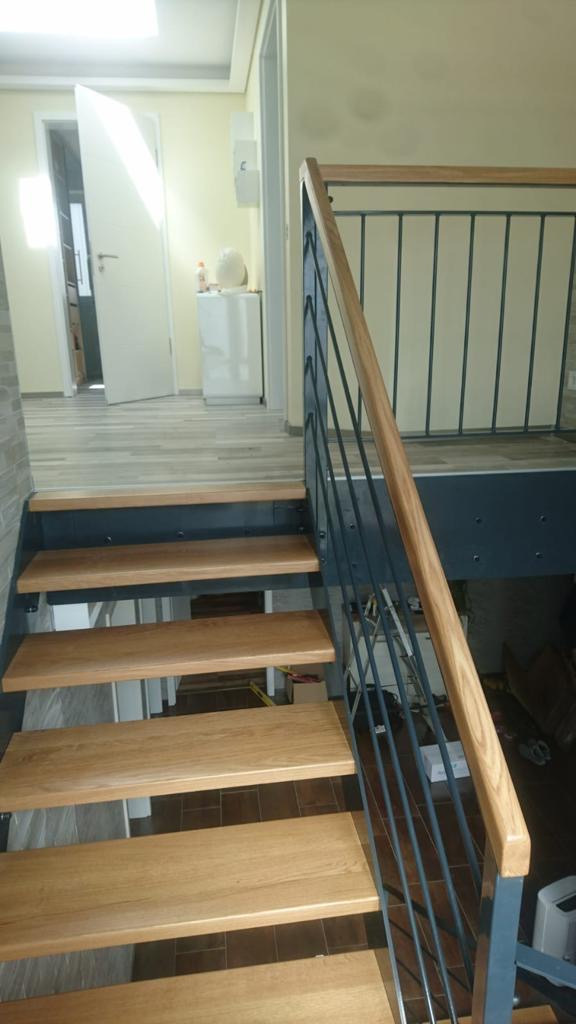 Drewniane schody z metalową balustradą o minimalistycznym designie, widok z dołu na górę, jasne wnętrze z otwartymi drzwiami w tle.