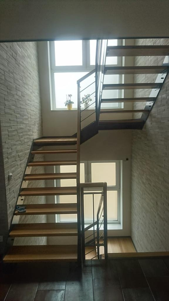 Nowoczesne schody z drewnianymi stopniami i metalową balustradą, oświetlone naturalnym światłem z okna, z widocznym fragmentem ściany wykończonej dekoracyjną cegłą.