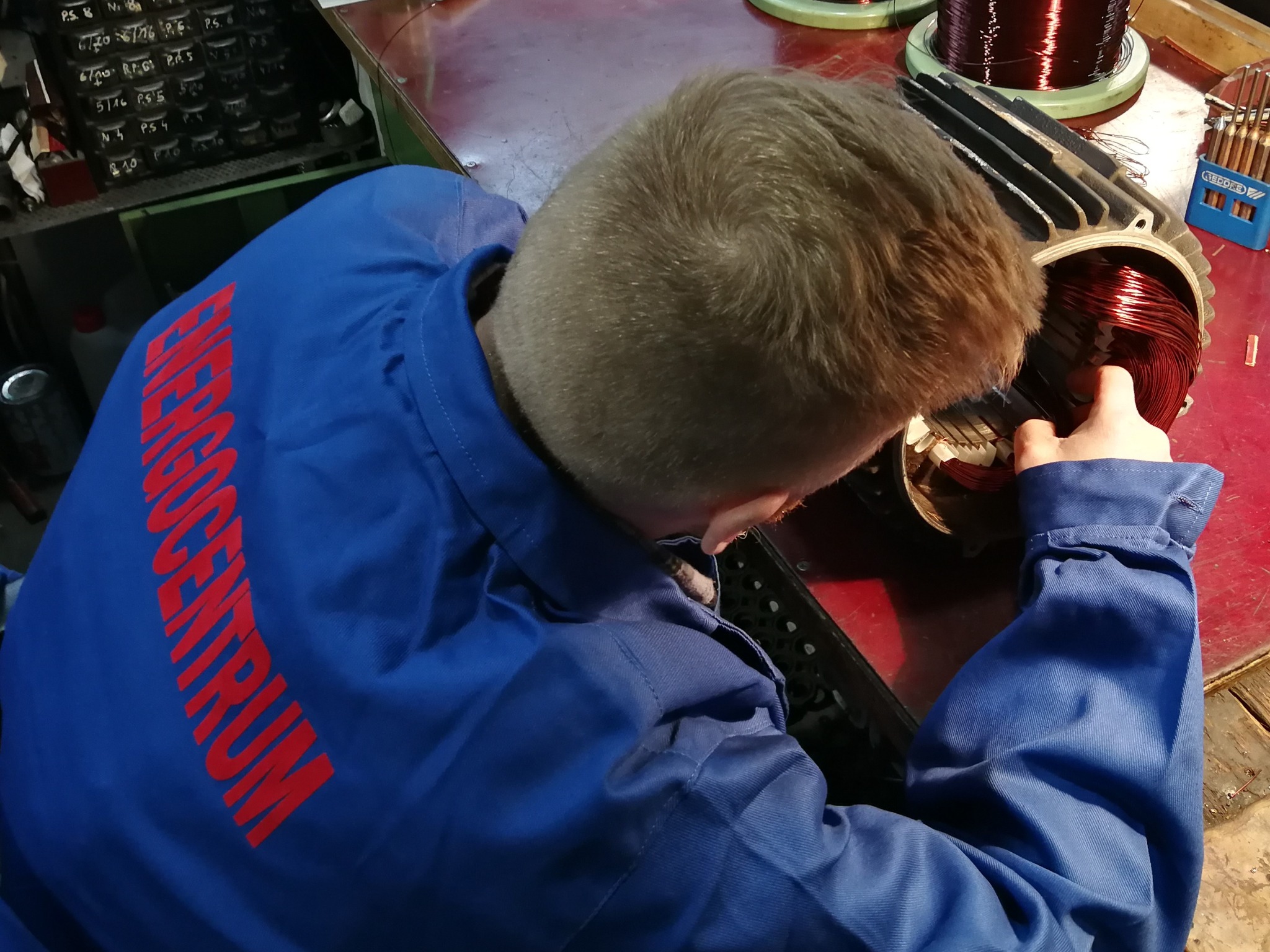 Elektromechanik w niebieskim kombinezonie z czerwonym napisem 'ENERGCENTRUM' nawija miedziane uzwojenie na stojan silnika elektrycznego na warsztatowym stole.