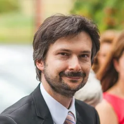 Portret uśmiechniętego mężczyzny w garniturze i krawacie w pastelowe paski, na rozmytym tle w plenerze.