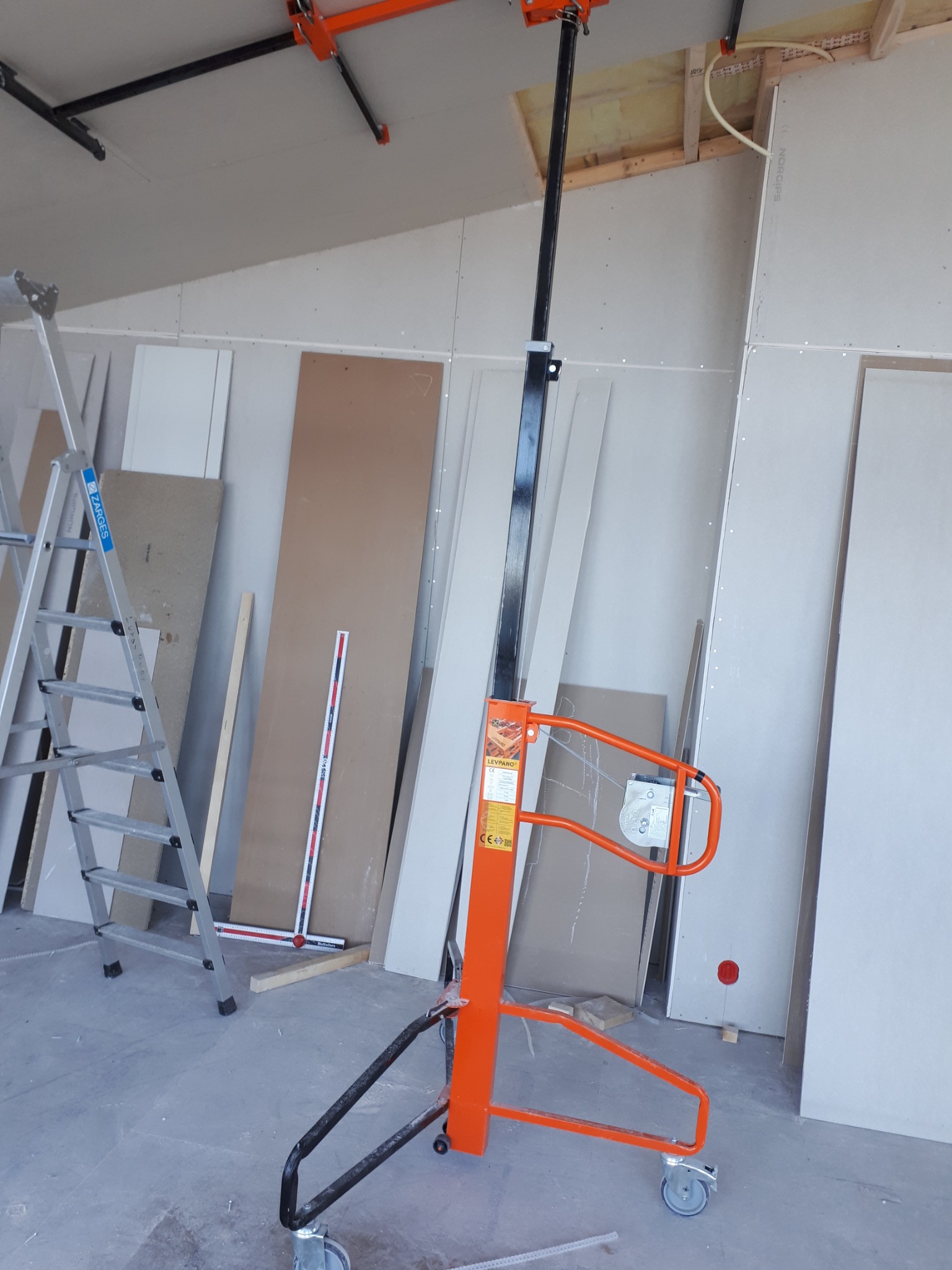 Pomarańczowy podnośnik do płyt kartonowo-gipsowych na tle ścian z zamontowanymi płytami, widoczna drabina aluminiowa i miarka.