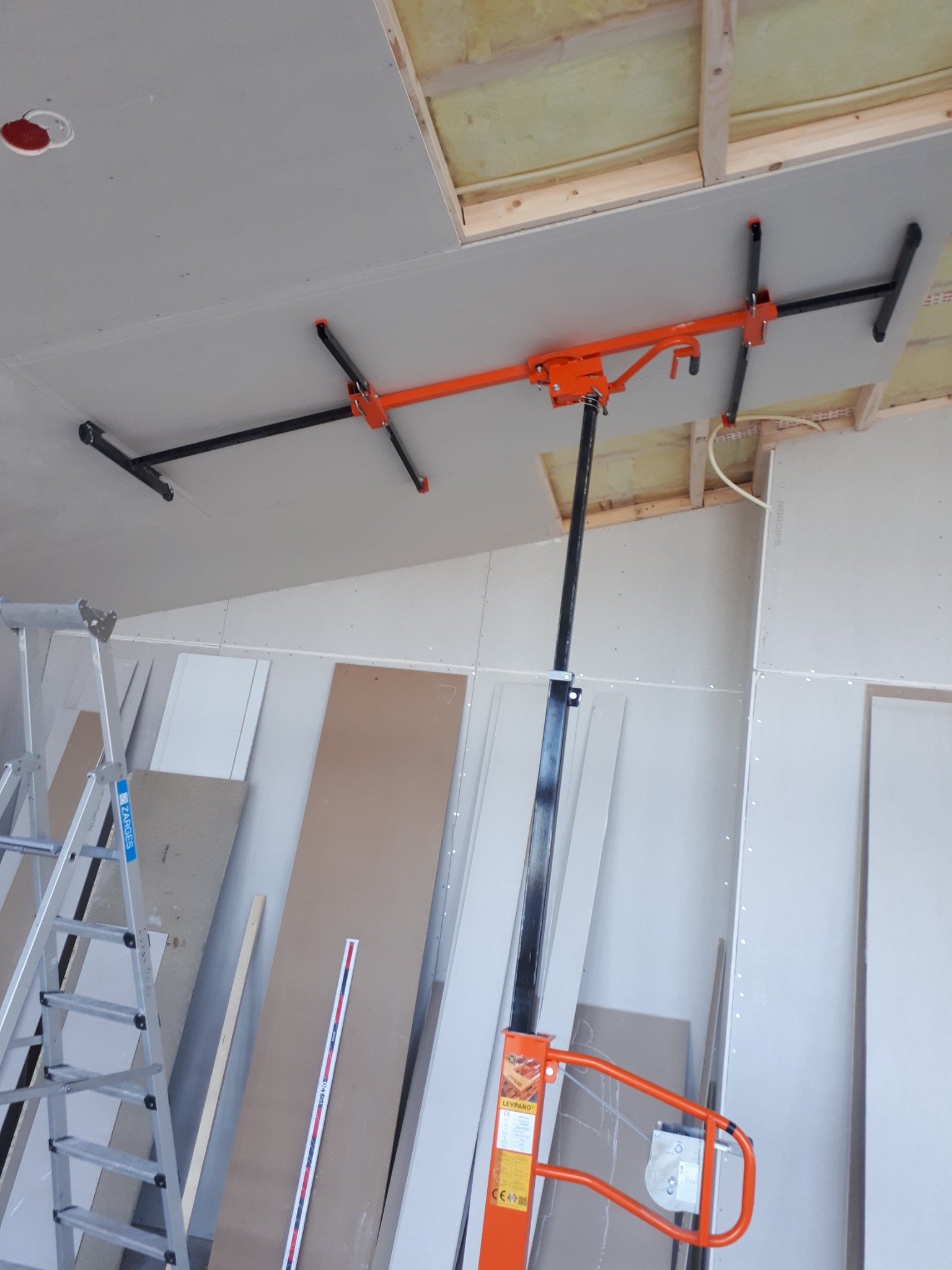 Montaż płyty gipsowo-kartonowej na suficie za pomocą podnośnika LEVPANO, widoczna drabina aluminiowa i płyty w tle.
