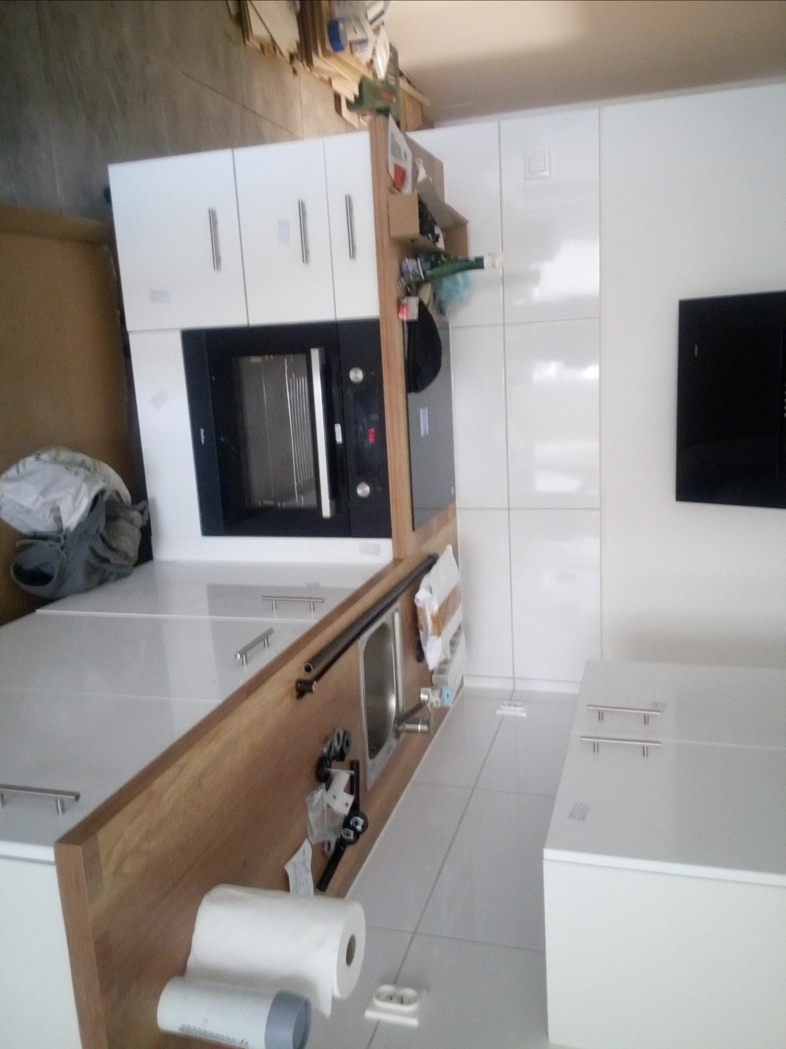 Nowoczesna biała kuchnia z drewnianym blatem, widoczne białe szafki z podłużnymi uchwytami, piekarnik w zabudowie, płyta grzewcza, zlewozmywak, na blacie leżą rolki papieru oraz elementy montażowe.