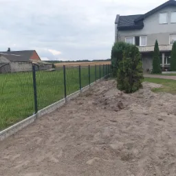 Nowo postawione, ciemne ogrodzenie panelowe z podmurówką z jasnej cegły na tle domu i budynków gospodarczych na wsi.