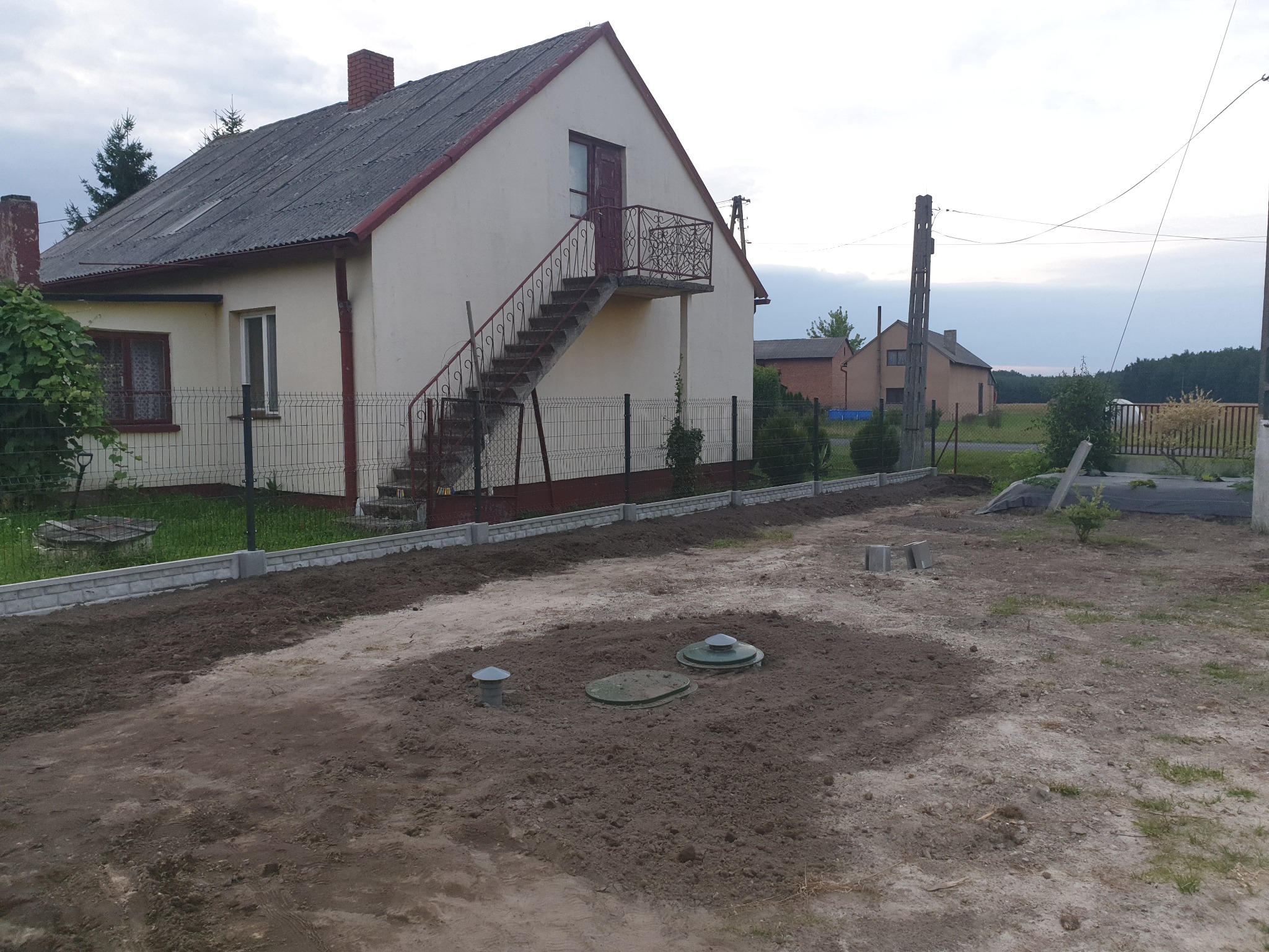 Nowo postawione ogrodzenie panelowe z podmurówką betonową otacza dom z zewnętrznymi schodami i balkonem, widoczne włazy do szamba na przygotowanym terenie.
