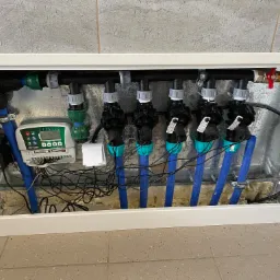 Rozdzielacz systemu nawadniania w metalowej skrzynce, z widocznym sterownikiem Rain Bird ESP-RZXe, zaworami elektromagnetycznymi, niebieskimi rurami i okablowaniem.