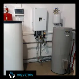 Kotłownia z zamontowanym białym modułem sterującym, szarym zbiornikiem Ferroli, czerwonym zbiornikiem ciśnieniowym, instalacją rurową i okablowaniem w pomieszczeniu z szarą podłogą.