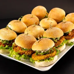 Rząd r&oacute;wno ułożonych mini burger&oacute;w z kotletami warzywnymi, sałatą, rukolą i sosem, spiętych wykałaczkami, na białej prostokątnej tacy na czarnym tle.