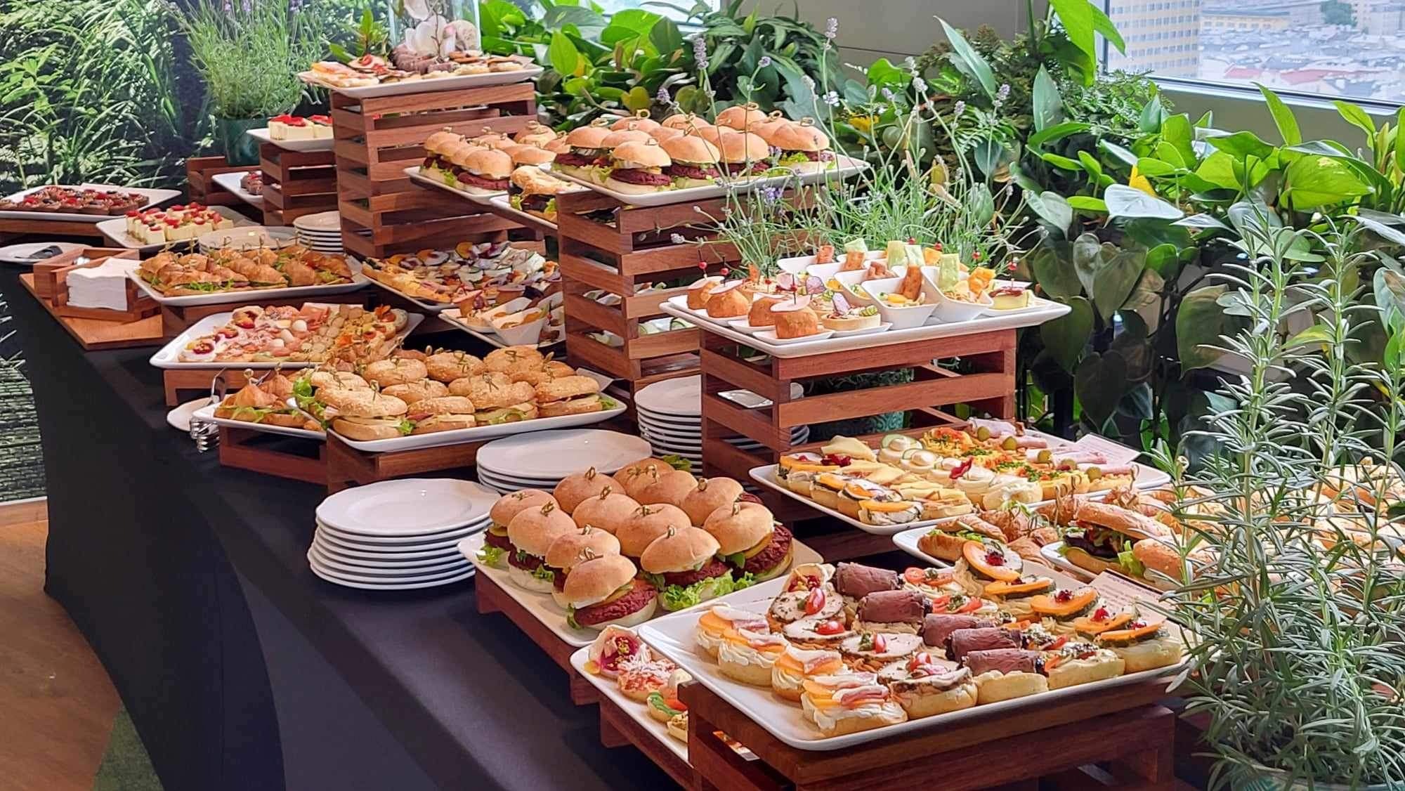 Bogato zastawiony stół cateringowy z różnorodnymi przekąskami, w tym mini burgerami, kanapeczkami, ciastami i sałatkami, na drewnianych podestach i białych talerzach, z zielonymi roślinami w tle...