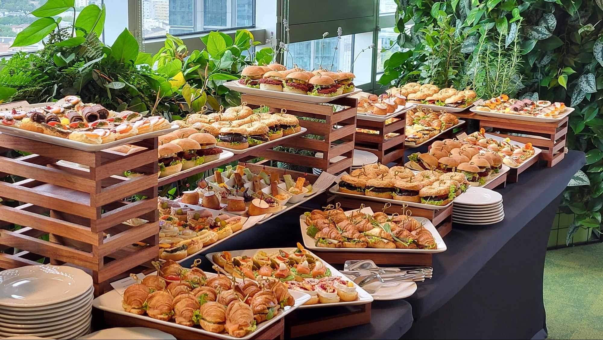 Bogato zastawiony stół z przekąskami na przyjęciu, z mini burgerami, roladkami z szynki, kanapeczkami i mini croissantami, ustawionymi na białych paterach i drewnianych podestach, z zielenią w tle.