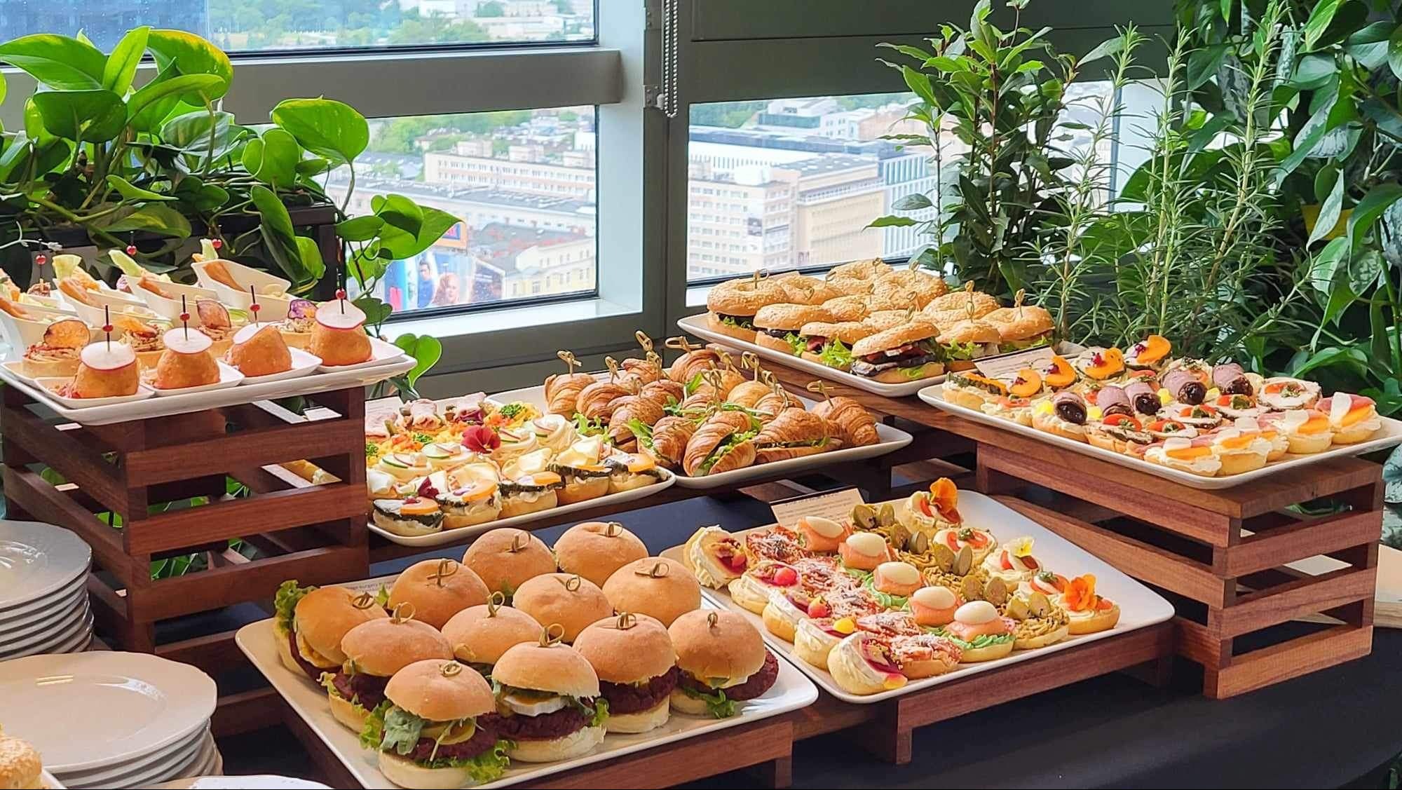 Elegancki bufet z różnorodnymi przekąskami: mini burgery, kanapki z croissantów, tartinki i koreczki na białych tacach, ustawione na drewnianych podestach, z zielonymi roślinami w tle i widokiem...