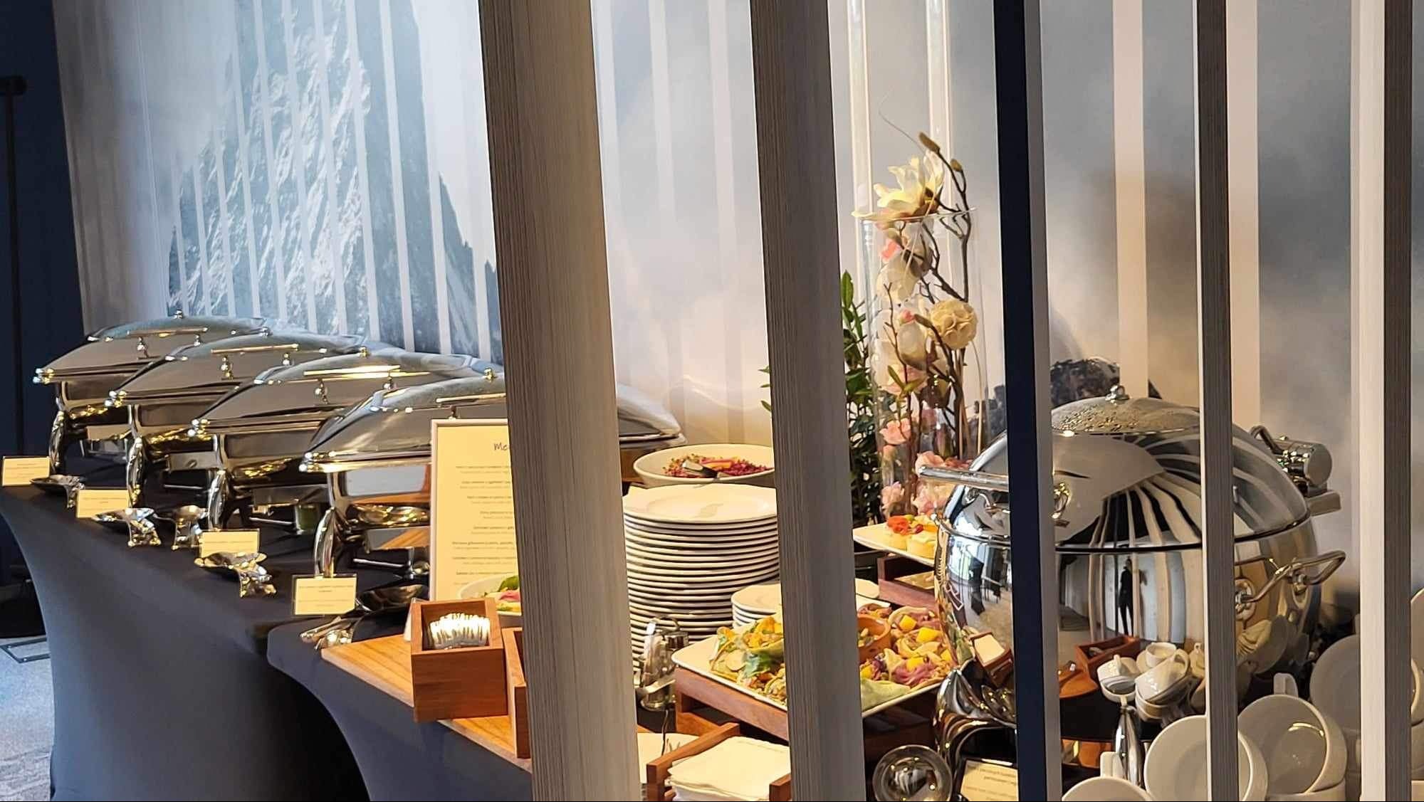 Elegancki stół cateringowy z podgrzewaczami, stosami talerzy, dekoracjami kwiatowymi i widocznym menu, widziany przez dekoracyjne przesłony.