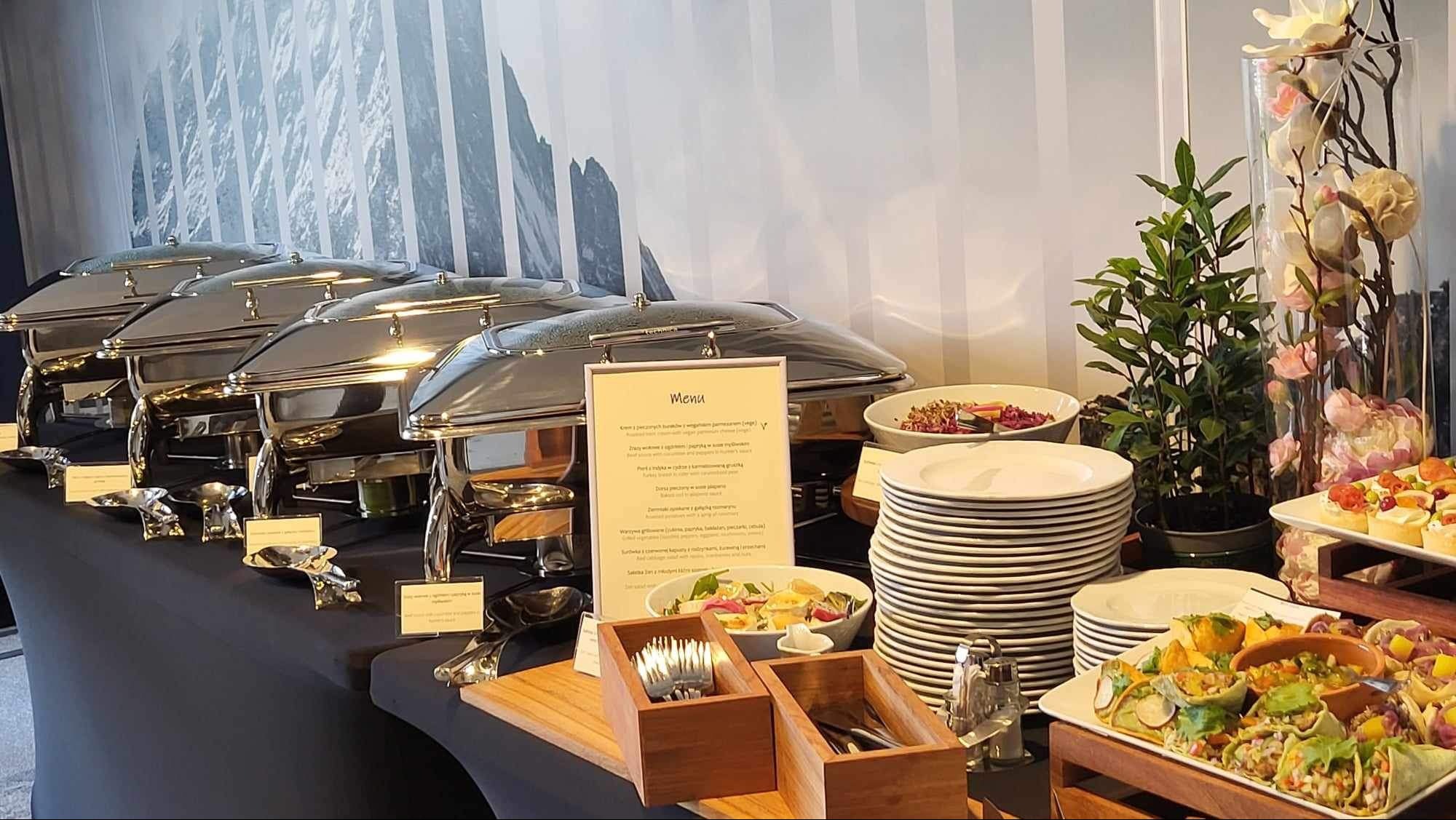 Ustawiony stół cateringowy z podgrzewaczami potraw, menu, stosami talerzy, sztućcami w drewnianych pojemnikach, sałatkami i przekąskami, dekoracjami roślinnymi na tle ściany z motywem górskim.