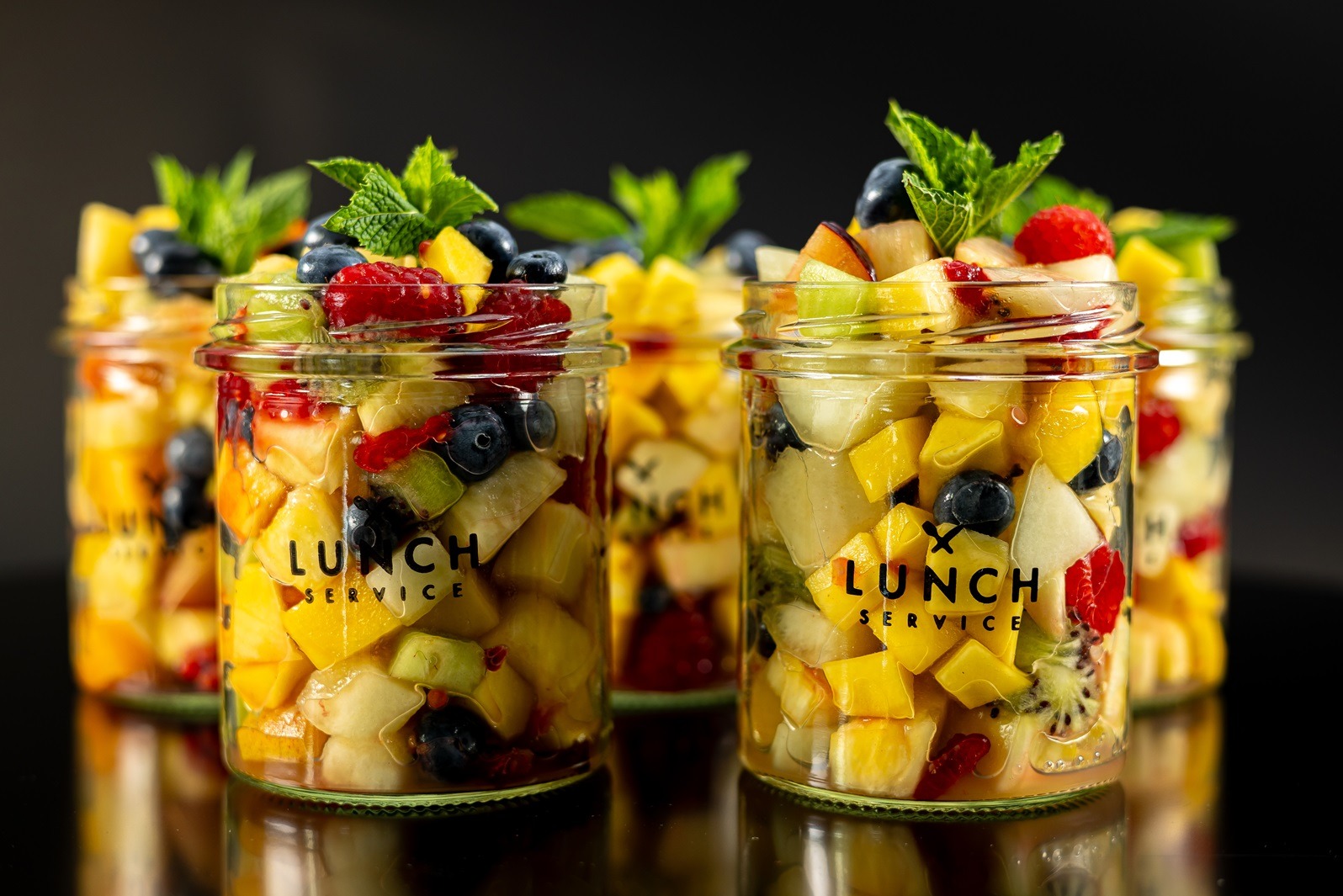 Szklane słoiki z pokrojonymi owocami (mango, kiwi, maliny, borówki) i listkami mięty, na każdym słoiku naklejka z napisem Lunch Service, ustawione na czarnym, błyszczącym tle.