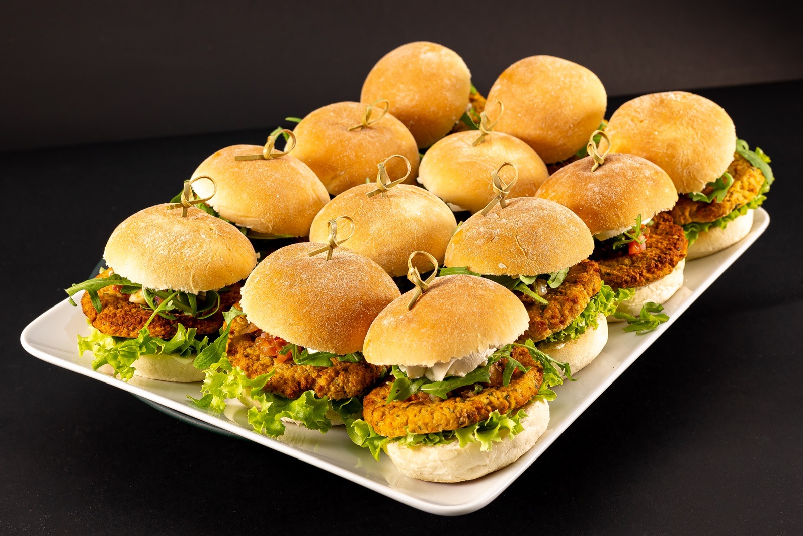 Rząd równo ułożonych mini burgerów z kotletami warzywnymi, sałatą, rukolą i sosem, spiętych wykałaczkami, na białej prostokątnej tacy na czarnym tle.
