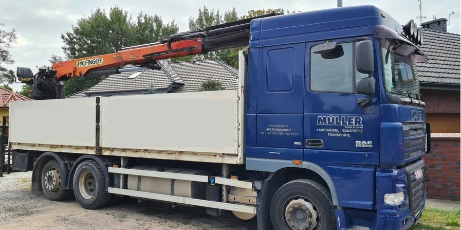 Niebieska ciężarówka DAF z pomarańczowym dźwigiem Palfinger na tle domów i drzew.