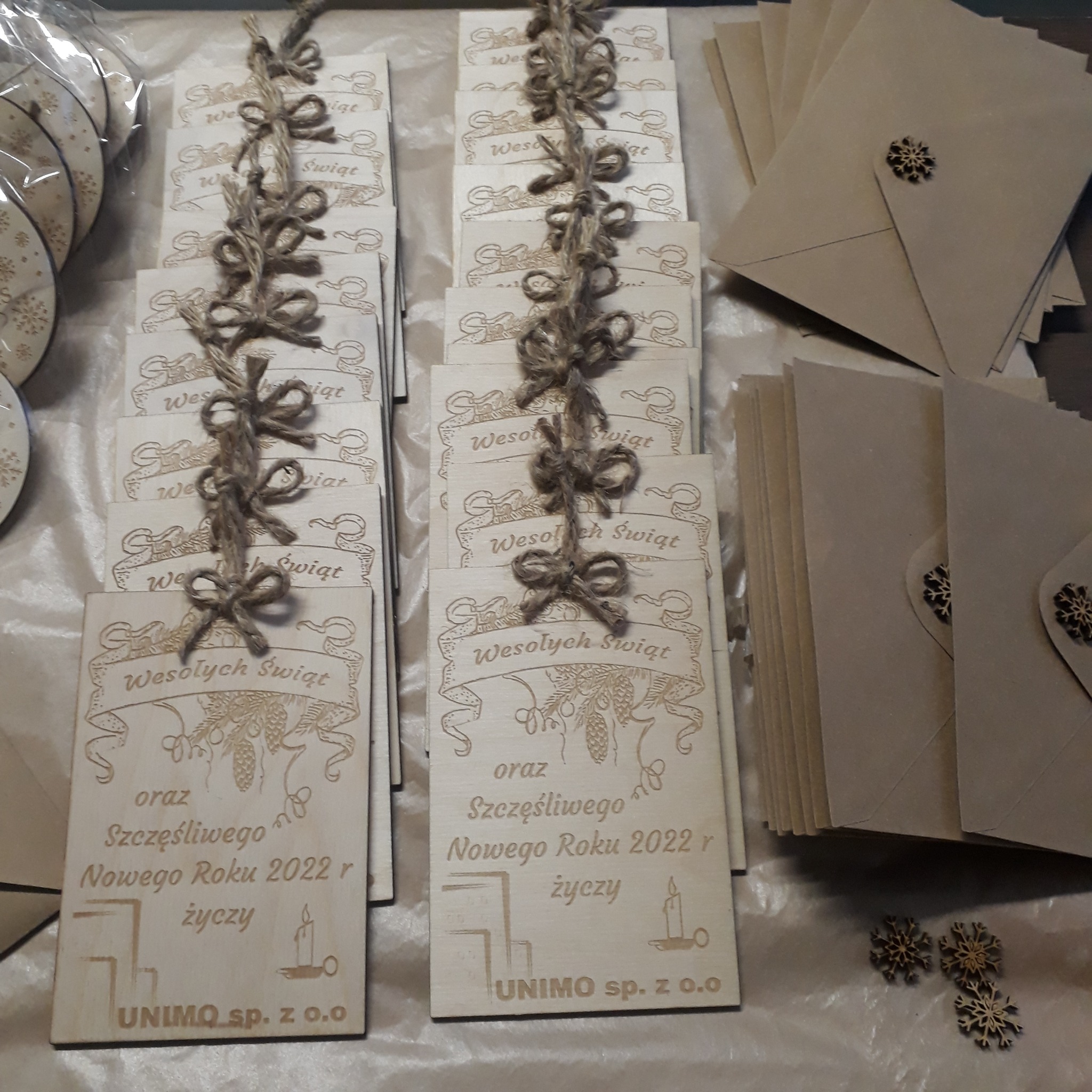 Ręcznie robione, drewniane kartki świąteczne z wygrawerowanym napisem 'Wesołych Świąt' oraz życzeniami noworocznymi, ozdobione sznurkiem jutowym i zapakowane w koperty z drewnianymi śnieżynkami.