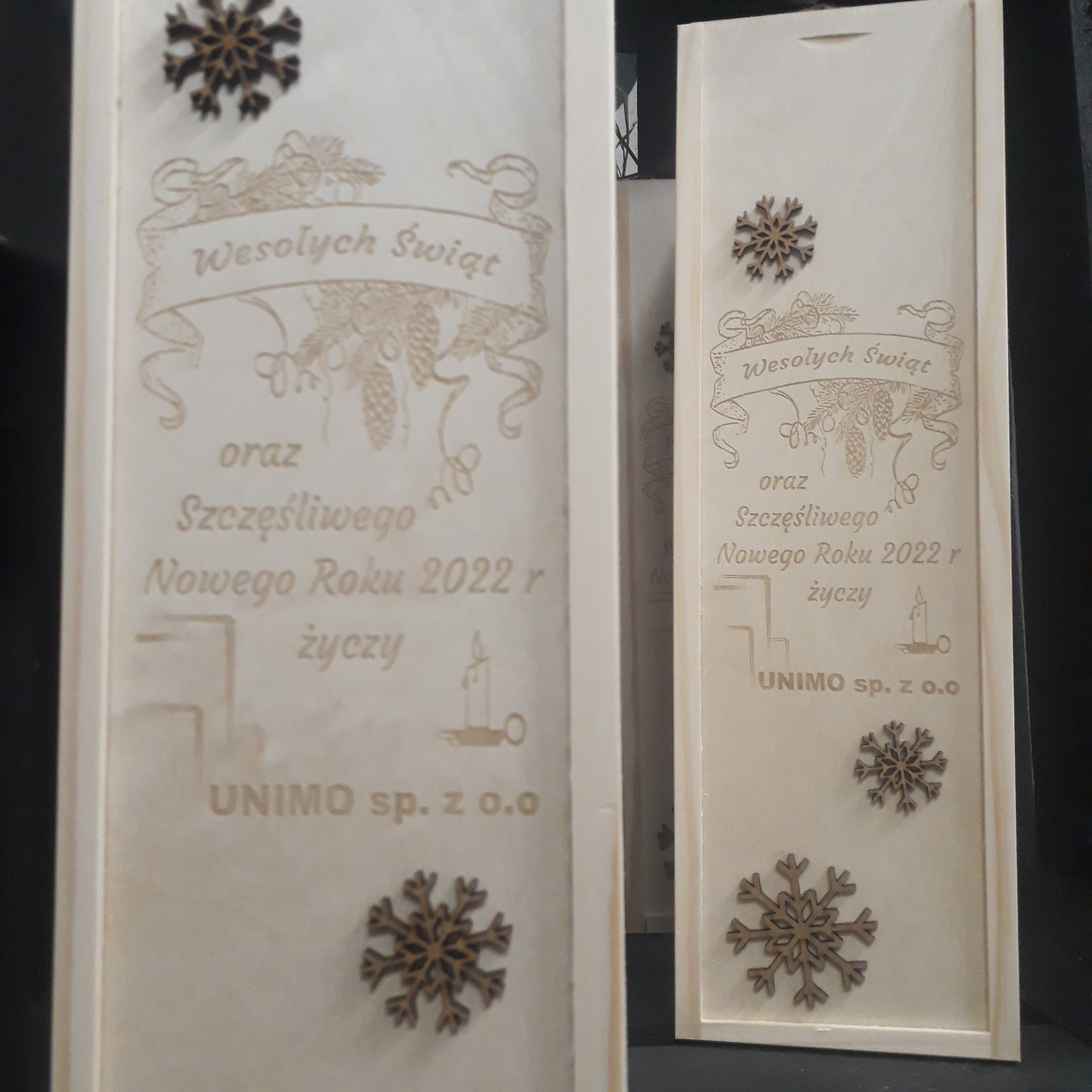 Dwa drewniane pudełka z wygrawerowanymi życzeniami 'Wesołych Świąt oraz Szczęśliwego Nowego Roku 2022 r' i nazwą firmy 'UNIMO sp. z o.o.', ozdobione drewnianymi śnieżynkami.