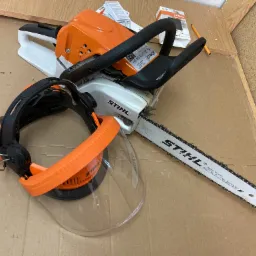 Piła spalinowa Stihl z osłoną twarzy i słuchawkami na kartonowym podłożu, obok instrukcja obsługi i ołówek.