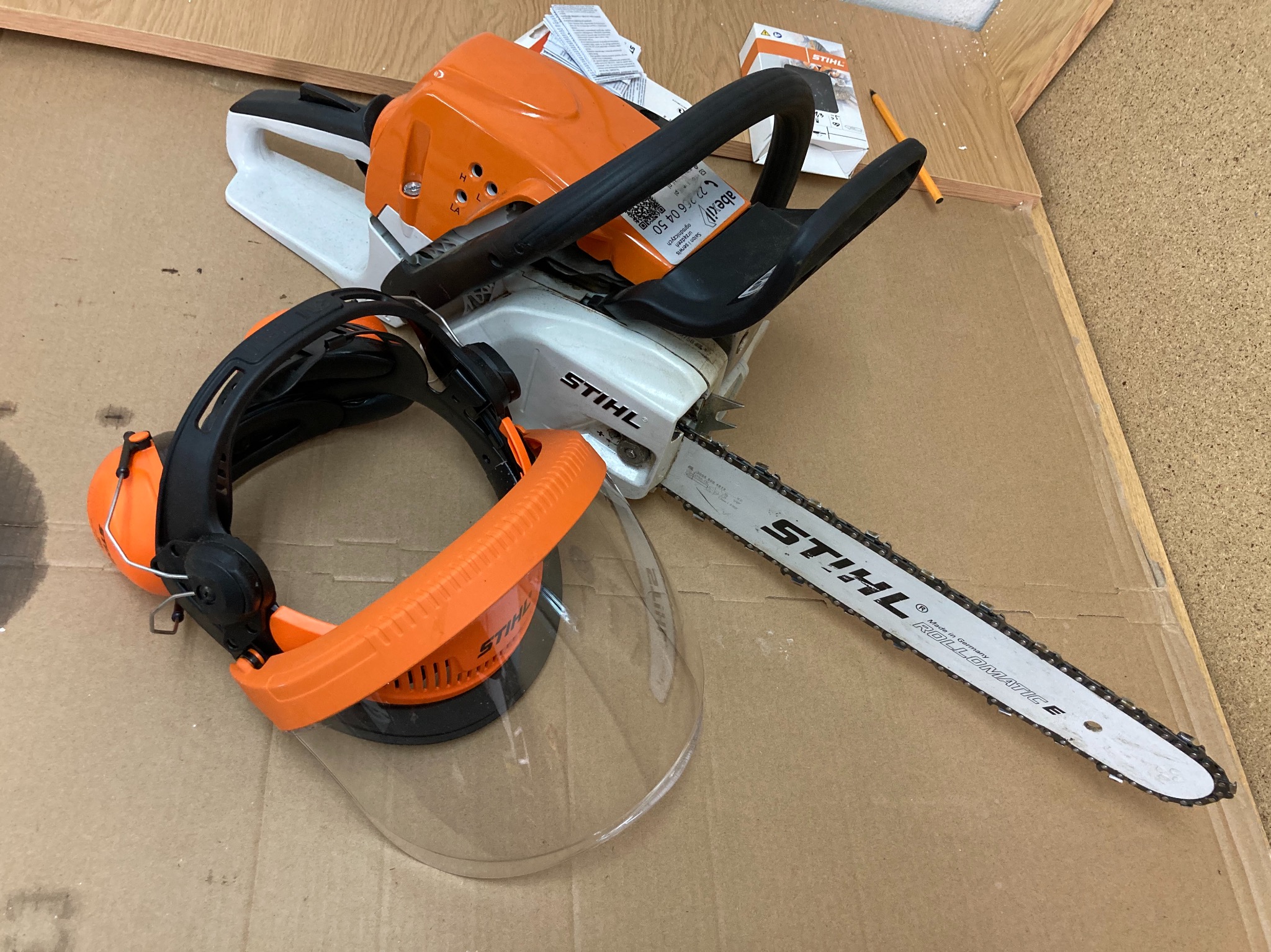 Piła spalinowa Stihl z osłoną twarzy i słuchawkami na kartonowym podłożu, obok instrukcja obsługi i ołówek.